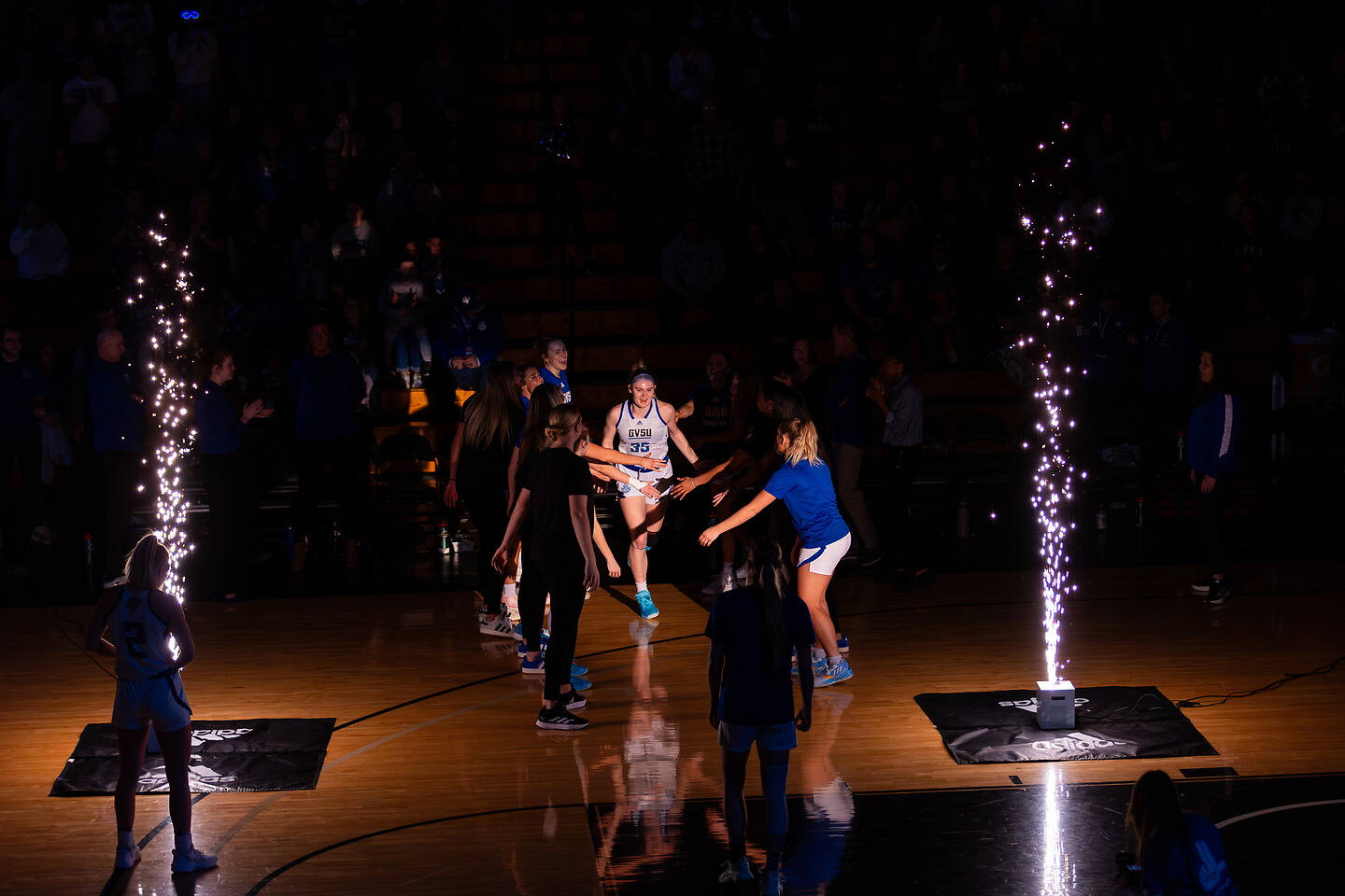 A basketball player in a white jersey is celebrated on the court as sparklers illuminate the scene, surrounded by teammates and staff.
